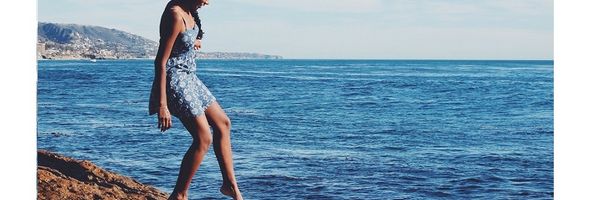 Unsicher, ob das Wasser zu kalt ist, hält eine junge Frau im Sommerkleid einen Fuß über das Meer.
